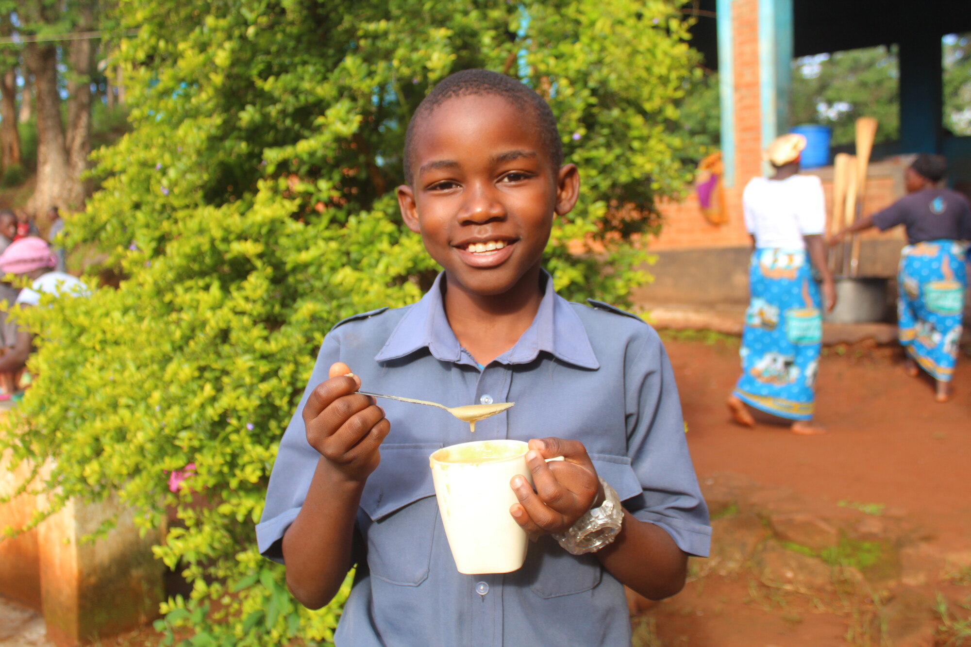 Steven, a young boy from Malawi, smiling to camera with a mug of porridge and a spoon