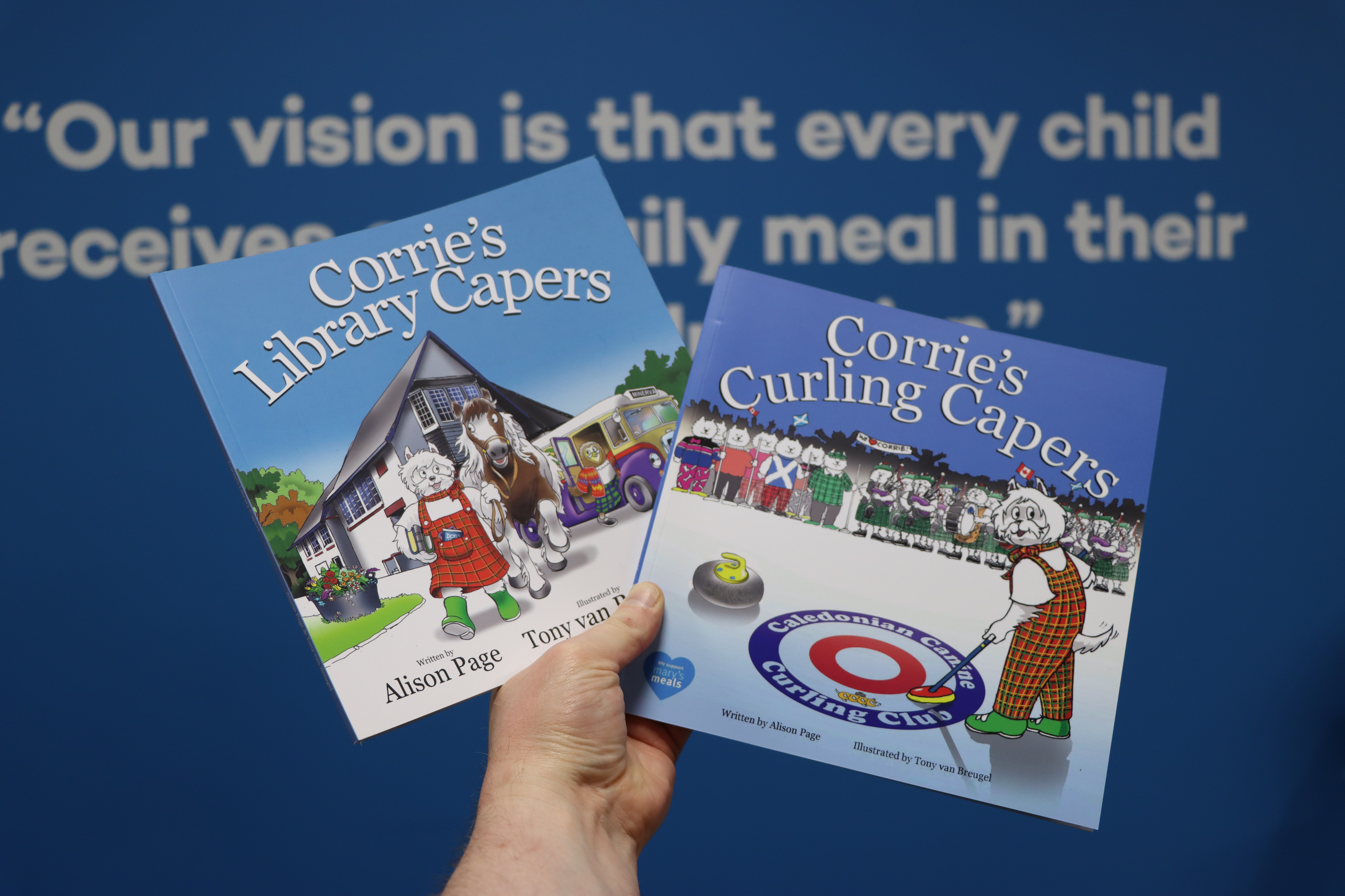 A hand holds up copies of two picture books, Corrie's Library Capers and Corrie's Paper Capers