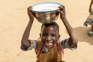 A young child from Benin carrying a silver bowl on his head and smiling at the camera