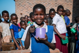 Smiling girl with mug and queue of children waiting to be served Mary's Meals behind her