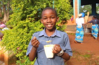 Steven, a young boy from Malawi, smiling to camera with a mug of porridge and a spoon