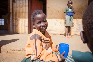 A child in Zambia sits outside smiling with a cup of Mary's Meals porridge in hand