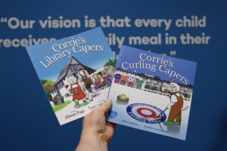 A hand holds up copies of two picture books, Corrie's Library Capers and Corrie's Paper Capers