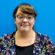 A woman with glasses and brown hair stands smiling against a blue wall 