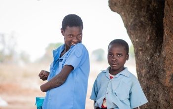 Fridah and Annette standing together next to a tree. Fridah is smiling and Annette is holding a mug