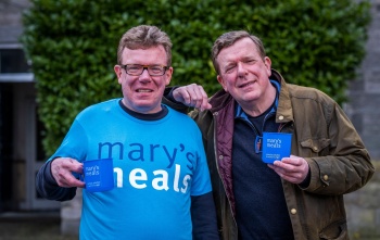 Charlie Reid stands on the left in a Mary's Meals t-shirt holding a mug and Craig Reid rests his arm on Charlie's shoulder and also holds a Mary's Meals mug.