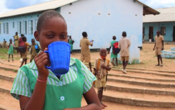 A girl in a green shirt with a blue cup covering her mouth, she is standing outside a school with children behind her