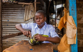 A teenager sits at a small desk repairing a pair of old shoes