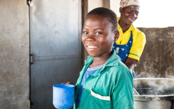 child in kitchen holding a mug, they are smiling. A woman cooks in a large stove behind the child.