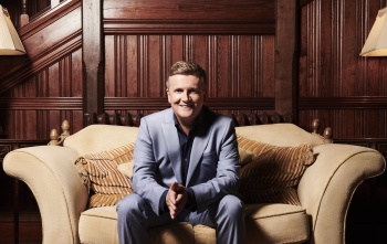 Aled Jones sitting on a sofa with his hands clasped and smiling