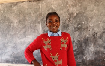 Anna wearing a red Christmas jumper with reindeer on, standing in front of a blackboard smiling
