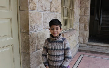 A young boy, Wissam, stands in a doorway smiling. He has dark hair and is wearing a striped top.