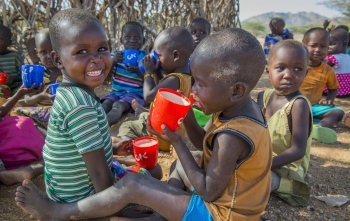 Two children, under 6, from Kenya sitting on the ground eating porridge from mugs