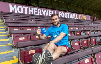 Football Stephen O'Donnell sitting in stand of football stadium in blue Mary's Meals t-shirt with blue Mary's Meals mug