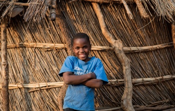 Boy stands in front of home with arms folded and smiling in Zambia