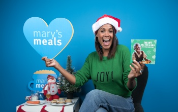 Jean Johansson sitting in a Santa hat, next to a Christmas table, holding up a Mary's Meals sign in one hand and a Christmas gift card in the other