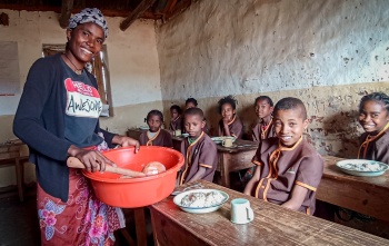 A volunteer cook serves food to children in a classroom
