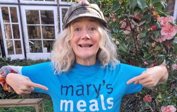 Actor Sophie Thompson stands outside wearing a Mary's Meals t-shirt, she is smiling and pointing at the Mary's Meals logo on the t-shirt
