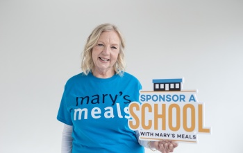 Deborah Meaden wearing a Mary's Meals t-shirt holding a sign which reads 'Sponsor A School with Mary's Meals'