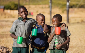 Three children with mugs of Mary's Meals porridge standing outside, smiling, in Zambia