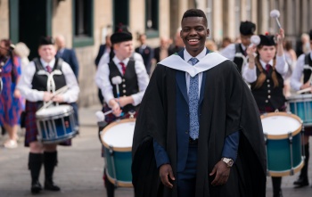 A smiling man in graduation gown stands in front of a Scottish pipe band