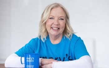 Deborah Meaden sitting smiling with her arms leaning on a table, next to a Mary's Meals mug