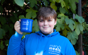 A young boy stands in a Mary's Meals hoodie holding a Mary's Meals mug