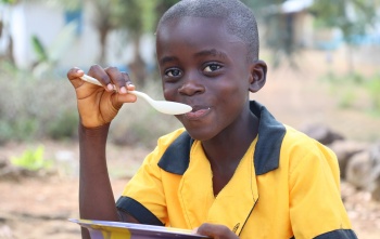A boy in yellow sits outside eating a bowl of food with a spoon