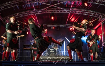 A group of men in kilts play bagpipes and guitars on a stage