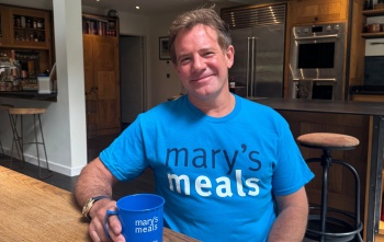 Matt Tebbutt sits at a kitchen table wearing a Mary's Meals t-shirt and holds a Mary's Meals mug
