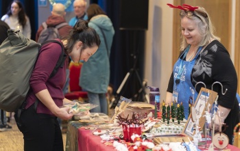 A woman looks at products at a stall at a Christmas market while another woman stands behind the stall