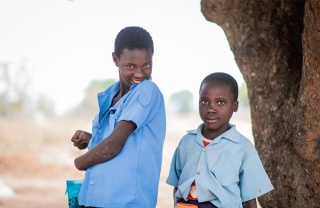 Fridah and Annette standing together next to a tree. Fridah is smiling and Annette is holding a mug