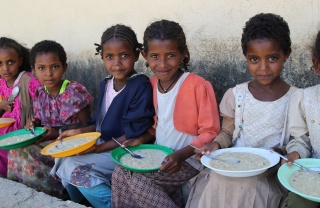 Children sitting on a wall eating bowls of food