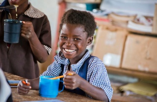 Smiling child sitting at a school desk eating Mary's Meals from a cup