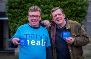 Charlie Reid stands on the left in a Mary's Meals t-shirt holding a mug and Craig Reid rests his arm on Charlie's shoulder and also holds a Mary's Meals mug.
