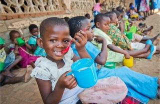 Children in Malawi eating from mugs while sitting on the ground together. The child closest to the camera is looking at it and smiling
