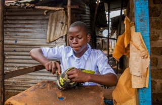 A teenager sits at a small desk repairing a pair of old shoes