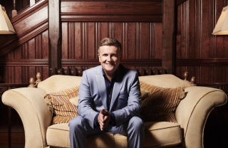 Aled Jones sitting on a sofa with his hands clasped and smiling