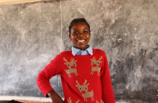 Anna wearing a red Christmas jumper with reindeer on, standing in front of a blackboard smiling