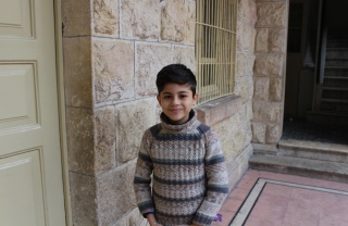 A young boy, Wissam, stands in a doorway smiling. He has dark hair and is wearing a striped top.