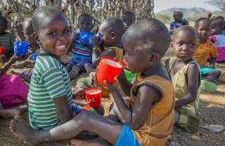 Two children, under 6, from Kenya sitting on the ground eating porridge from mugs