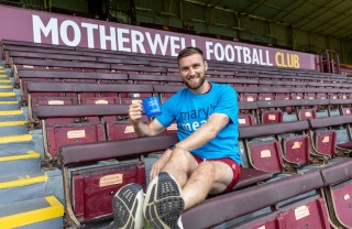 Football Stephen O'Donnell sitting in stand of football stadium in blue Mary's Meals t-shirt with blue Mary's Meals mug