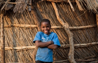Boy stands in front of home with arms folded and smiling in Zambia