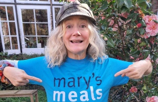 Actor Sophie Thompson stands outside wearing a Mary's Meals t-shirt, she is smiling and pointing at the Mary's Meals logo on the t-shirt