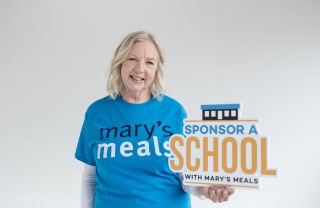 Deborah Meaden wearing a Mary's Meals t-shirt holding a sign which reads 'Sponsor A School with Mary's Meals'