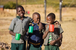Three children with mugs of Mary's Meals porridge standing outside, smiling, in Zambia