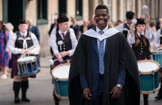 A smiling man in graduation gown stands in front of a Scottish pipe band