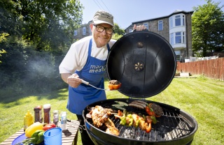 Gary Maclean holds a piece of meat over a BBQ with food cooking