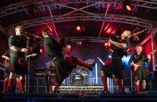 A group of men in kilts play bagpipes and guitars on a stage