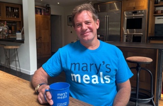 Matt Tebbutt sits at a kitchen table wearing a Mary's Meals t-shirt and holds a Mary's Meals mug